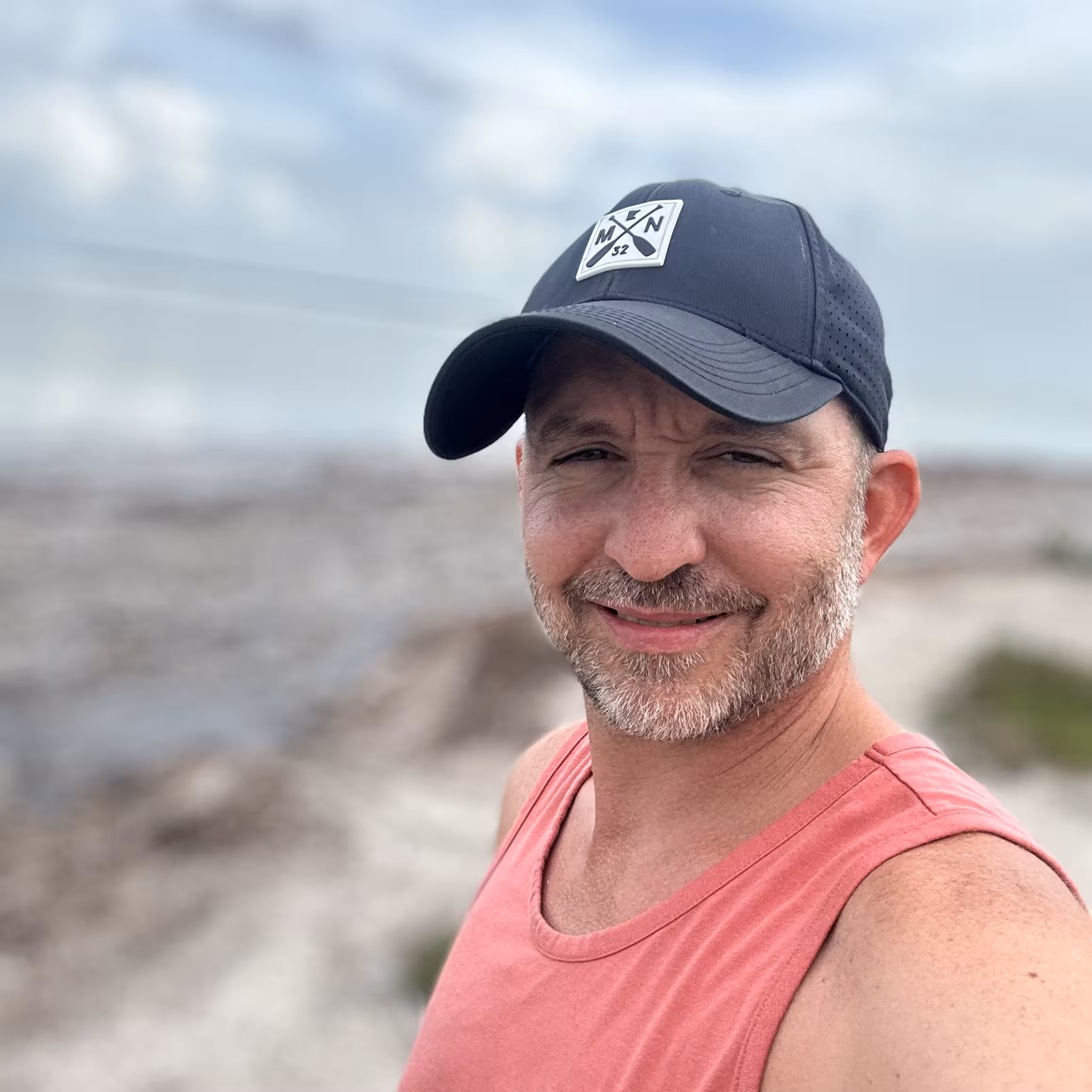 White man in black cap and coral tank top smiling by rocky shoreline under a cloudy sky.
