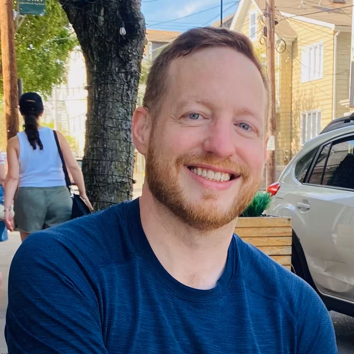 White man with short red hair and beard, smiling in a navy shirt outdoors near a tree and parked car.