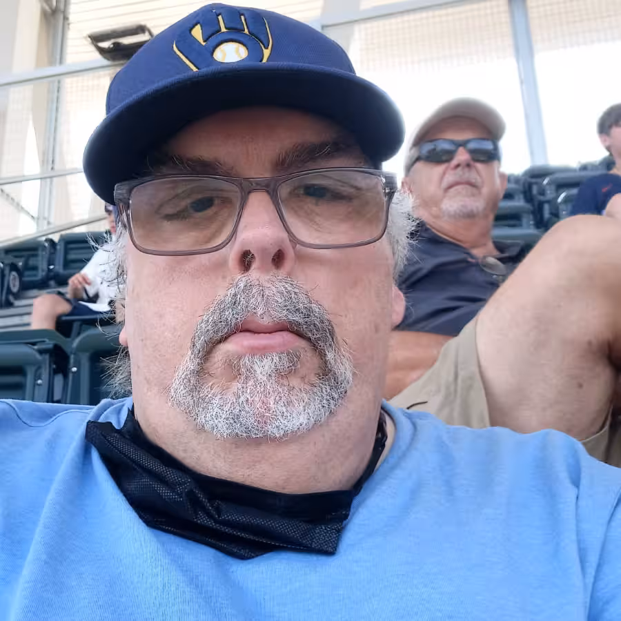 White man with gray mustache and goatee, glasses, dark cap, and light blue shirt sitting in stadium seats.
